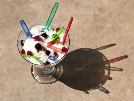 Glass bowl of frozen yogurt with fresh berries and many colorful plastic spoons in sunlight