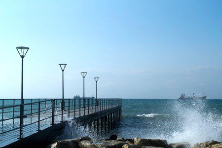 Empty sea pier with street lamps, splashing waves and the light blue sky