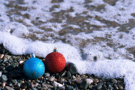 Two colorful Christmas balls at the beach among sea pebbles near foamy waveの写真素材