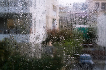 Wet window glass with rain drops and blurred silhouettes of houses, trees and car