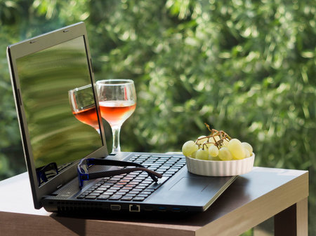 Laptop, eyeglasses, glass of wine and bowl of grapes on garden table