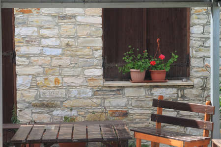Brown wooden table and empty bench in front of old stone wall with shuttered window and two flower potsの写真素材