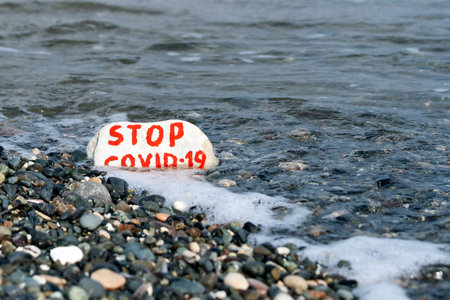 White stone with written message Stop COVID-19 laying in sea waves on the beach
