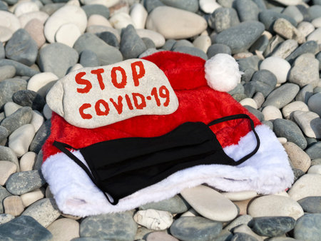 Santa hat, protective face mask and stone with message Stop COVID-19 on beach pebbles in sunlight