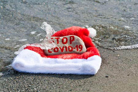 Santa hat and stone with message Stop COVID-19 in sea wave on sandy beachの写真素材