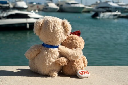 Two teddy bears in love sitting in harbor, hugging each other and looking at the sea with yachts and...