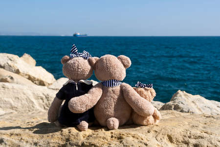 Teddy bear family sitting on big rock in front of blue sea and sky with ship on the horizonの写真素材