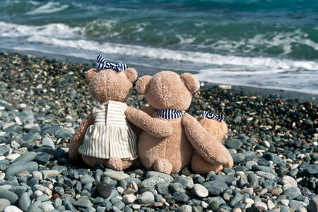 Family of three teddy bears sitting on stone beach in front of wavy seaの写真素材