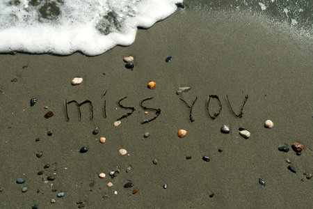 Words Miss You written on wet sand among pebbles, seashells beside foamy wave on sea beach