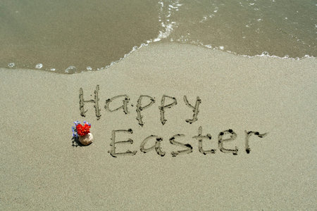 Phrase Happy Easter written on wet beach sand beside sea wave