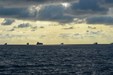 Several cargo ships are silhouetted against a dramatic sky with dark clouds. The calm ocean features...
