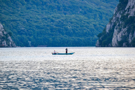 A lone fisherman stands on a boat in the Danube River, surrounded by forested hills and rocky cliffs near the Iron Gates Gorge, Romania and Serbia.の写真素材