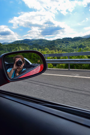 View from inside a car on a rural road, focusing on a side mirror reflecting a person with a camera....