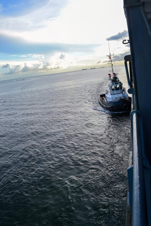 A tugboat moves through calm waters beside a larger vessel. The sky is partly cloudy, and the sun...