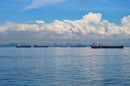 Large cargo ships float on calm waters with city skyline in the background. Fluffy clouds fill the sky, adding depth to the tranquil maritime scene.の写真素材