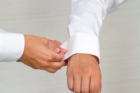A detailed view of one person assisting another in securing a cufflink on a white dress shirt, with...