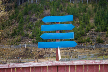 A wooden signpost with four blank blue arrows stands near a rustic fence, set against a forested hillside in a rural area with sparse vegetation.の写真素材