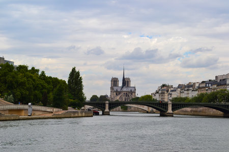 Notre Dame Cathedral's twin towers and spire rise in the background, with a bridge crossing the Seine River, tree lined embankment, and Parisian buildings.の写真素材
