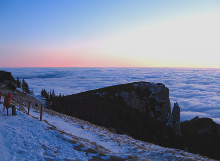 A snow covered trail leads to a wooden fence as a person stands nearby. A rocky outcrop and evergreen trees frame a sea of clouds at sunset.の写真素材