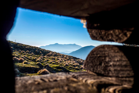 A scenic view of rolling grassy hills and a jagged mountain ridge, framed by wooden logs. Soft blue haze suggests early morning or late afternoon light.の写真素材