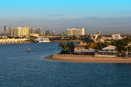 A coastal city features a sandy beach with palm trees, calm blue waters, boats, a large ferry, a bridge, and a skyline of mid rise and high rise buildings.の写真素材