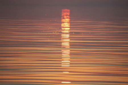 Horizontal lines of orange and purple water ripples reflect a golden sun streak at dusk, creating a serene and tranquil atmosphere.の素材