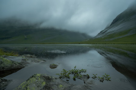 A moody mountain landscape with a lake reflecting misty clouds. Raindrops create ripples on the dark water, with green hills and rocks in the foreground.の素材