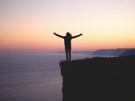 A solitary figure stands silhouetted on a cliff edge, arms wide, facing a serene ocean at sunset.の素材