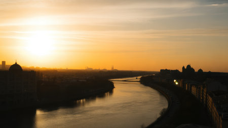 A tranquil cityscape at sunset with a river reflecting the warm orange and yellow hues of the sky. Domed buildings and a bridge are silhouetted along the shore, creating a serene and peaceful...の素材