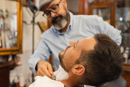 Barber shaving a man's beard with a barber's razor over shaving soap applied to the neck with a brush in the barbershop.の写真素材