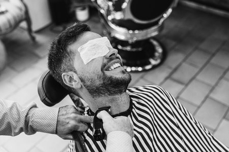 Black and white photo of a barber trimming the beard with an electric razor of a man with eye protection sitting in a barber chair in a barbershop.の写真素材