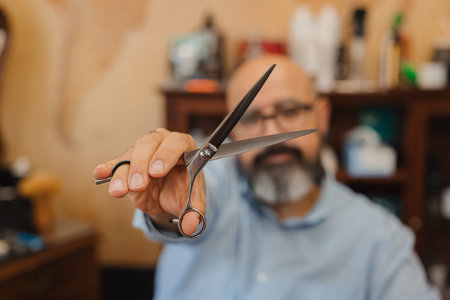 A man holding a pair of scissors in his hand. He is in a barber shop. The scissors are black and...