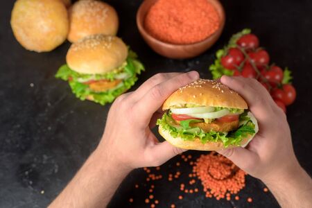 vegetarian hamburger with lentil cutlet, avocado, tomatoes, onions, lettuce on a wooden table on a dark background. delicious Burger for vegetarians. Burger in the hands of a man close up.の写真素材