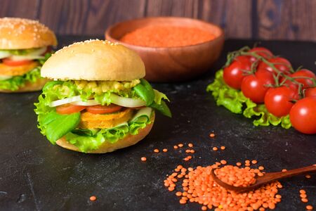 vegetarian hamburger with lentil cutlet, avocado, tomatoes, onions, lettuce on a wooden table on a dark background. delicious Burger for vegetarians. close up. lentil seeds are scatteredの写真素材