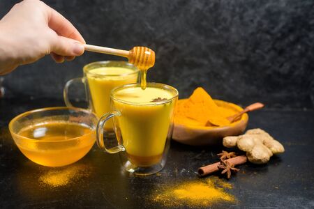Indian traditional Golden milk with turmeric, ginger, spices, honey. add a spoonful of honey to the milk. healing effect of the drink. ingredients for a Golden drink on a dark backgroundの写真素材
