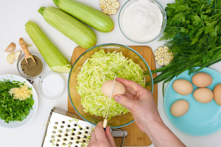 step-by-step recipe to cook pancakes of zucchini and greens on a light background. with the addition of eggs, flour, garlic, pepper. break eggs to grated zucchini in a plate. the view from the top.の写真素材