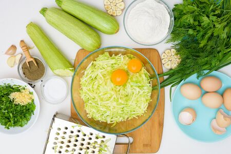 step-by-step recipe to cook pancakes of zucchini and greens on a light background. with the addition of eggs, flour, garlic, pepper. break eggs to grated zucchini in a plate. the view from the top.の写真素材