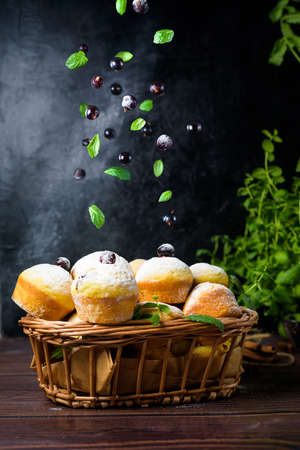 flying cupcakes with mint leaves and black currants in powdered sugar on a black background, a wooden basket with cupcakes and black currants . levitation of pancakes and muffins.の写真素材