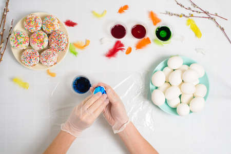 the process of coloring eggs for Easter in blue and red colors. a woman in gloves paints eggs with dye from a jar, top view, willow branches and colorful feathers decor.の写真素材