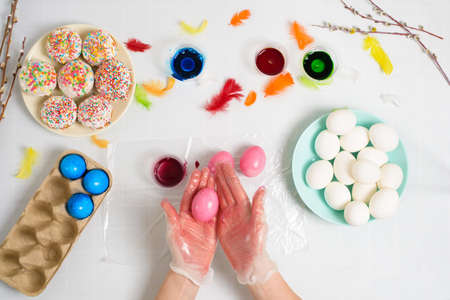the process of coloring eggs for Easter in blue and red colors. a woman in gloves paints eggs with dye from a jar, top view, willow branches and colorful feathers decor.の写真素材