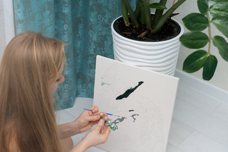 hobbies at home self-isolation, painting by numbers to draw from the top view of a woman holding a brush with paints, painting. the home interior is light.のeditorial素材