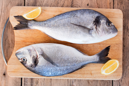 Two fresh gilt-head bream fish with slices of lemon on cutting board on wooden の写真素材