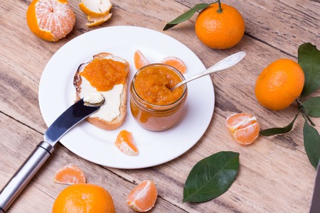 toasted slice of bread with butter and homemade tangerine jam on white plate. Colorful tangerine jam in glass jar on rustic wooden boardの写真素材