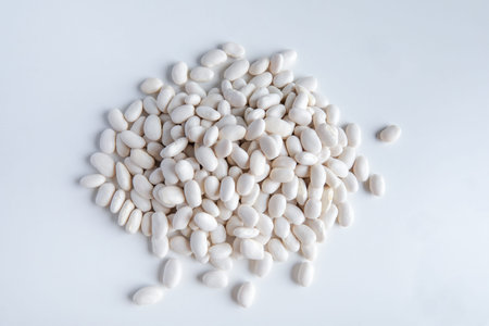 White beans on a white background, top view, close-up. A pile of small, oval white beans is...
