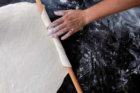 A top-down view of a hand rolling out dough with a wooden rolling pin on a dark, floured surface.