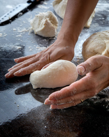 A close-up of hands shaping a piece of dough into a small loaf on a dark floured countertop. Several...