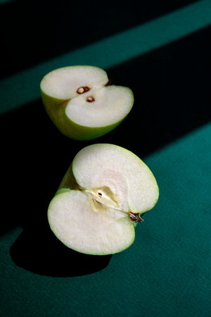 Minimalist composition of a fresh green apple cut in half, placed on a deep green surface with...