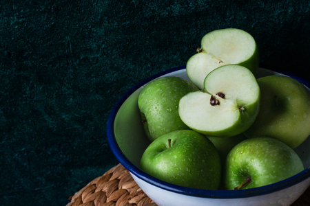 Close-up of fresh green apples in a white enamel bowl with blue rim, placed on a rustic mat against a dark textured background.の写真素材