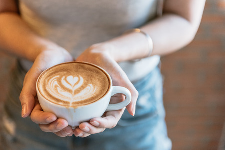 Female hands holding a cup of coffee with foam over wooden table, top view vintage tone.の写真素材
