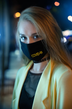 Portrait of young stylish woman looking in camera in protective mask at night.